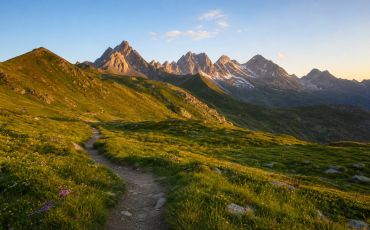 Plan large d'un alpage en été, sommets encore un peu enneigés en arrière-plan, sentier de randonnée qui serpente, lumière chaude de matinée ou fin d'après-midi. 